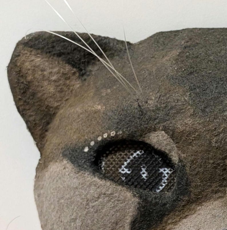May include: Close-up of a felted cat head with a detailed eye and whiskers. The cat's fur is a mix of gray and brown, with a darker shade around the eye. The eye is black with white stitching, and the whiskers are thin and silver.