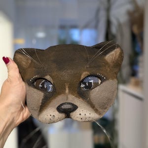 May include: A brown cat mask with detailed facial features, including mesh eyes and whiskers. The mask is hand-painted with a realistic design, showcasing a playful and artistic aesthetic. This unique mask is suitable for costume parties or theatrical performances.