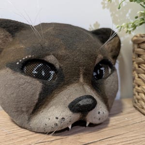May include: A handcrafted, brown and gray cat mask with mesh eye coverings. The mask features a black nose, white whiskers, and small teeth. The mask is made of a felt-like material and is designed to be worn.