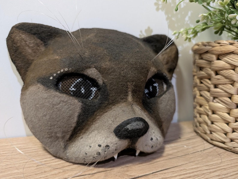 May include: A handcrafted, brown and gray cat mask with mesh eye coverings. The mask features a black nose, white whiskers, and small teeth. The mask is made of a felt-like material and is designed to be worn.