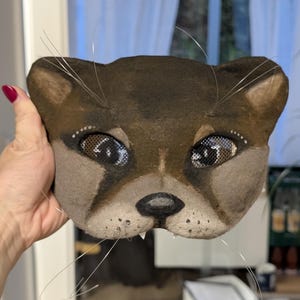 May include: A handcrafted, brown and tan cat mask with mesh eyes and wire whiskers. The mask features a detailed design with a dark brown nose, small white teeth, and a light brown muzzle. The cat mask is a unique piece of art.