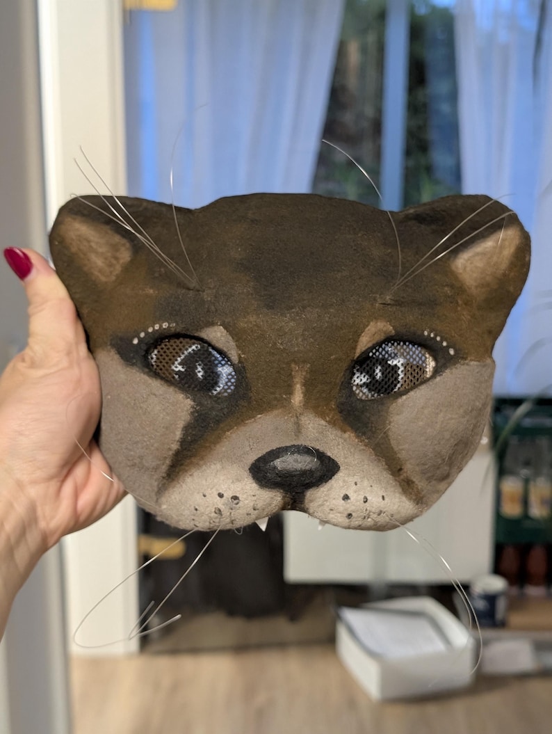 May include: A handcrafted, brown and tan cat mask with mesh eyes and wire whiskers. The mask features a detailed design with a dark brown nose, small white teeth, and a light brown muzzle. The cat mask is a unique piece of art.