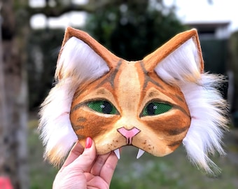 May include: A handmade orange and white cat mask with green eyes and fangs. The mask is made of felt and fur and has a realistic look.