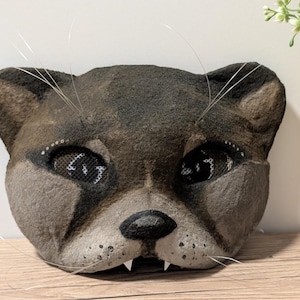 May include: A handmade cat mask with a textured, gray and brown surface. The mask features detailed eyes with mesh coverings, a black nose, and white whiskers. The cat mask has small, white fangs.