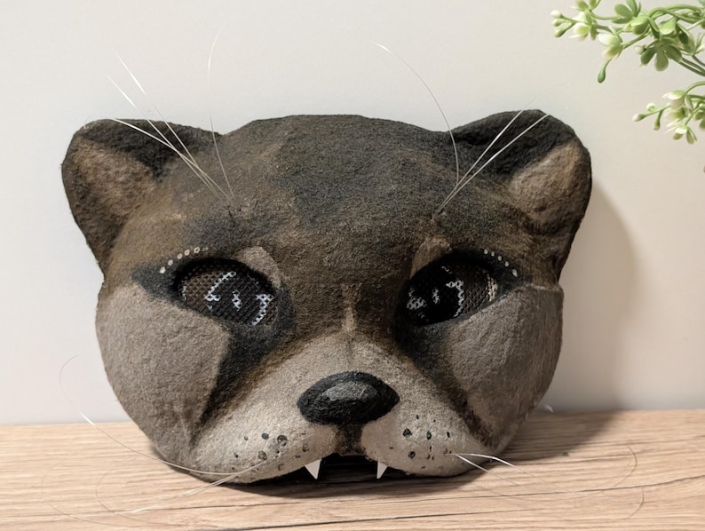 May include: A handmade cat mask with a textured, gray and brown surface. The mask features detailed eyes with mesh coverings, a black nose, and white whiskers. The cat mask has small, white fangs.