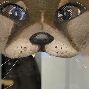 May include: A close-up of a brown and tan cat mask with mesh eyes and whiskers. The mask has a black nose, white teeth, and small white dots above the eyes. The mask is held by a hand.
