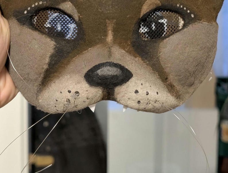 May include: A close-up of a brown and tan cat mask with mesh eyes and whiskers. The mask has a black nose, white teeth, and small white dots above the eyes. The mask is held by a hand.