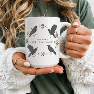 May include: A white ceramic mug featuring a circular graphic of four black crows, small mushrooms, and botanical elements. The text "SUPPORT YOUR LOCAL MURDER" is printed in the center in a simple black font. The mug has a large handle.