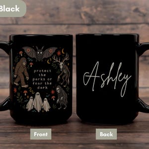 May include: A solid black ceramic mug featuring a graphic illustration of a bat, a sasquatch, a deer-skulled creature, and three ghosts with the text "protect the parks or fear the dark" on the front and the name "Ashley" in white script on the back.