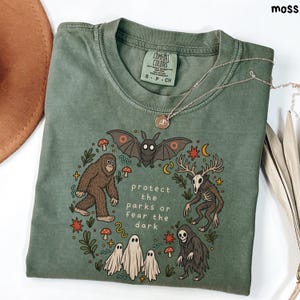 May include: A moss green t-shirt featuring a graphic print of a bat, a sasquatch, a deer skull creature, ghosts, and a grim reaper arranged in a circle. The text reads protect the parks or fear the dark. The shirt has a Comfort Colors brand label.
