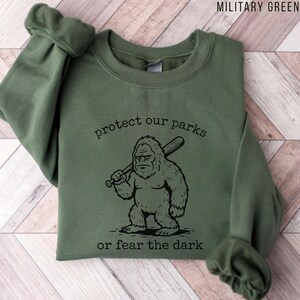 May include: A military green crewneck sweatshirt featuring a black graphic of a Bigfoot creature holding a baseball bat. The text above the creature reads protect our parks and the text below reads or fear the dark in a simple black font.