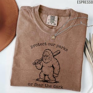 May include: An espresso-colored t-shirt featuring a graphic of a creature holding a baseball bat with the text "protect our parks" above and "or fear the dark" below. The shirt has a Comfort Colors brand label inside the neckline.