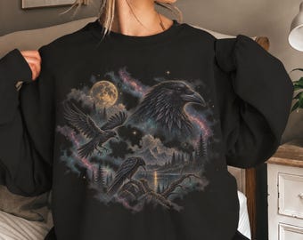 Peut inclure: Un sweat-shirt noir à col rond orné d'une impression graphique représentant deux corbeaux dans un ciel nocturne. Le motif comprend une grande pleine lune, un fond de galaxie nébuleuse, des montagnes, des sapins et un lac avec reflet.