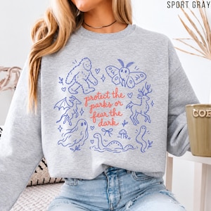 May include: A sport gray crewneck sweatshirt featuring a blue graphic print of a sasquatch, moth, dragon, ghost, deer, and sea creature. The text in the center reads, "protect the parks or fear the dark" in a red cursive font.
