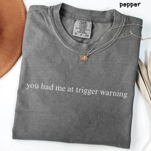 May include: A gray Comfort Colors t-shirt with the phrase "you had me at trigger warning" printed in white. A gold-colored necklace with a pendant is on the shirt. The shirt is folded and displayed on a white surface.