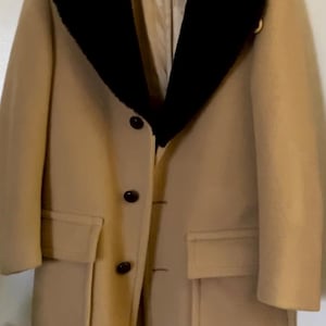 May include: A tan overcoat with a black collar and three dark buttons. The coat has two front pockets and a tailored fit. The coat is hanging on a white hanger against a white wall.