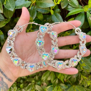 May include: Silver hoop earrings featuring heart-shaped iridescent crystals and clear rhinestones. The earrings are linked by a chain design, creating a glamorous accessory. The earrings are displayed against a backdrop of green foliage.