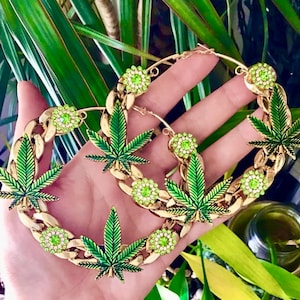 Può includere: Orecchini a cerchio color oro con disegni di foglie di cannabis verdi e accenti verdi scintillanti. Gli orecchini hanno uno stile a catena e sono impreziositi da pietre preziose verdi rotonde. Le foglie sono dettagliate con smalto verde.