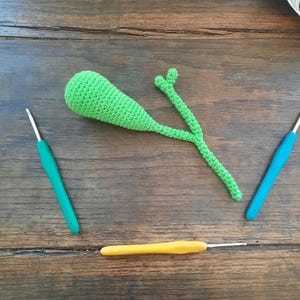 May include: A green crocheted amigurumi toy shaped like a pancreas. The toy is lying on a wooden surface with two crochet hooks, one green and one yellow.