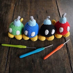Puede incluir: Cuatro juguetes submarinos de crochet en verde, azul, negro y rojo con detalles amarillos. Los juguetes están alineados en una superficie de madera con tres ganchos de crochet.