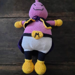 May include: A crocheted plush toy of a purple and pink character with a black belt and the letter "M" on it. The toy has yellow feet and is lying on a wooden surface.
