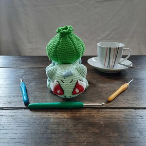 May include: A green crocheted Bulbasaur Pokemon plushie with red eyes. The plushie is sitting on a wooden table with two crochet hooks and a teacup and saucer.