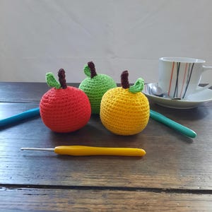 May include: Three crocheted apples in red, green, and yellow with brown stems and green leaves. The apples are sitting on a wooden table with a yellow crochet hook and a blue crochet hook.