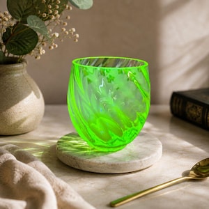 Puede incluir: Un vaso de vidrio verde neón vibrante con un patrón de remolinos marmolados en todo el material. El recipiente tiene una silueta redondeada sin tallo con un borde liso y pulido, y una calidad translúcida que permite el paso de la luz.