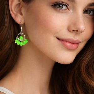Pode incluir: Um close-up de uma pessoa usando brincos de lustre verde brilhante. Os brincos têm um aro prateado com uma única conta verde no topo e várias contas verdes em forma de gota penduradas. A pessoa tem pele clara e cabelo castanho.