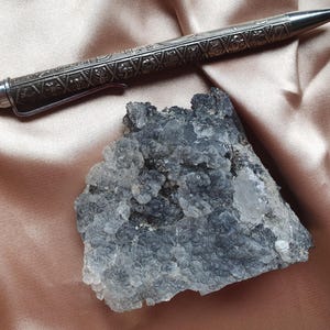 Op de afbeelding: Een zilverkleurige pen met gedetailleerde bloemen- en geometrische patronen ligt boven een cluster van blauwgrijze mineraalkristallen. De pen heeft een clip en een punt. Het mineraal heeft een ruw, getextureerd oppervlak.