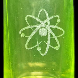 Può includere: Un bicchierino verde neon con un simbolo di atomo inciso bianco. Il bicchiere è cilindrico con una base spessa e un bordo leggermente svasato. Il simbolo dell'atomo è centrato.