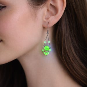 Op de afbeelding: Een zilveren oorbel met een hangend ontwerp. Het heeft een centrale, ronde, neon groene steen omringd door kleinere groene kralen. De kralen zijn verbonden door delicate kettingen, waardoor een krooneffect ontstaat. De oorbel wordt op het oor gedragen.