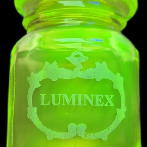 May include: A vivid neon green glass jar with a rounded body and a textured rim. The jar features a white, etched design with the word "LUMINEX". The jar is set against a black background, highlighting its bright colour.