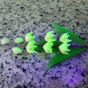 May include: A delicate glass lily of the valley flower brooch with green leaves. The flowers are a pale green color and are arranged in a cluster. The brooch is made of glass and wire.