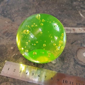 Puede incluir: Una esfera de vidrio verde llena de pequeñas burbujas de vidrio amarillo. La esfera está sobre una superficie gris texturizada.