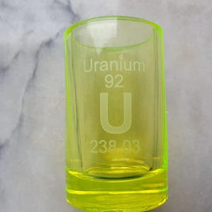 May include: A bright yellow shot glass with the text "Uranium 92 U 238.03" etched on the side.