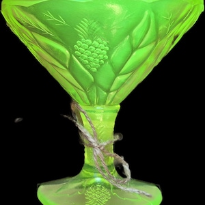 Puede incluir: Una copa de postre de vidrio verde neón vibrante con un motivo de piña moldeado en el cuenco y la base. El tallo está envuelto con un trozo de cuerda marrón natural. El vidrio tiene una calidad translúcida y un borde ancho y festoneado.