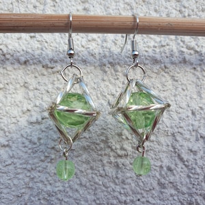 Könnte beinhalten: Ein Paar silberne Ohrringe mit geometrischem Design. Jeder Ohrring hat eine grüne Glasperle, die in einem Drahtkäfig hängt. Die Ohrringe haben unten eine baumelnde grüne Glasperle.