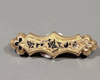 May include: A gold-toned brooch with a black enamel floral design. The brooch is shaped like a rectangle with rounded edges.