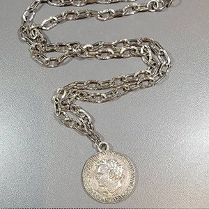 May include: Silver-toned necklace featuring a chain link design and a circular pendant. The pendant displays a profile portrait and Roman numerals. The necklace is laid out on a gray surface.