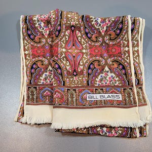 May include: A folded scarf with a paisley pattern in red, pink, and gold. The scarf has a cream-colored border with fringe and the text "BILL BLASS" on a label.