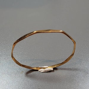 May include: A gold-colored, geometric bangle bracelet. The bracelet has an irregular, faceted design. The metal has a slightly hammered texture. The bracelet is resting on a small piece of white material against a gray background.