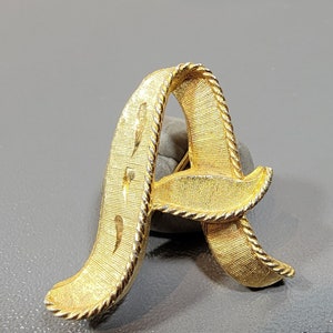 May include: A gold-toned letter A brooch with a rope-like design. The letter is stylized and has a curved shape.