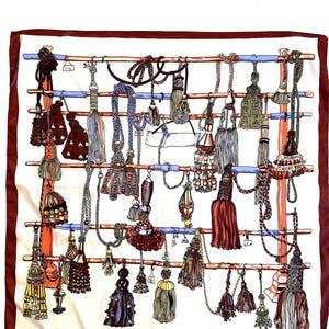May include: A white silk scarf with a burgundy border, featuring a detailed illustration of various decorative tassels. The tassels are in shades of red, brown, gold, and blue, hanging from a wooden structure with blue and orange accents.
