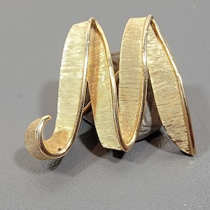 May include: A gold-toned metal brooch in the shape of the letter M. The letter is made of a textured, ribbon-like material.