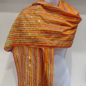 May include: A striped scarf with sequins in shades of orange, yellow, and pink. The scarf is draped over a white mannequin.