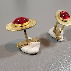 May include: A pair of gold cufflinks with red glass stones set in the center of each cufflink. The cufflinks are on a white surface.