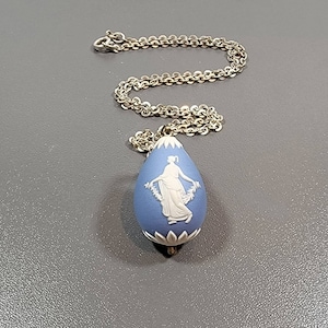 May include: A blue pendant necklace with a silver chain. The pendant is teardrop-shaped with a white bas-relief of a woman holding a garland. The pendant has a white floral design at the top and bottom.