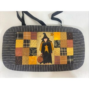 Könnte beinhalten: Eine rechteckige Halloween-Tasche mit einem schwarz-weiß gestreiften Rand. Die Mitte zeigt ein Patchwork-Design mit einer Hexenillustration, die einen Kürbis-Laterne hält. Die Hexe trägt einen schwarzen Hut und Umhang mit einem gelben Stern.