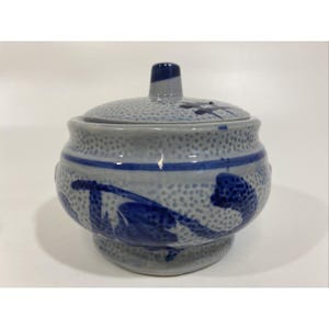 May include: A ceramic jar with a lid, featuring a speckled gray base and lid, adorned with blue floral designs and a blue stripe. The jar has a small, cylindrical handle on top of the lid.